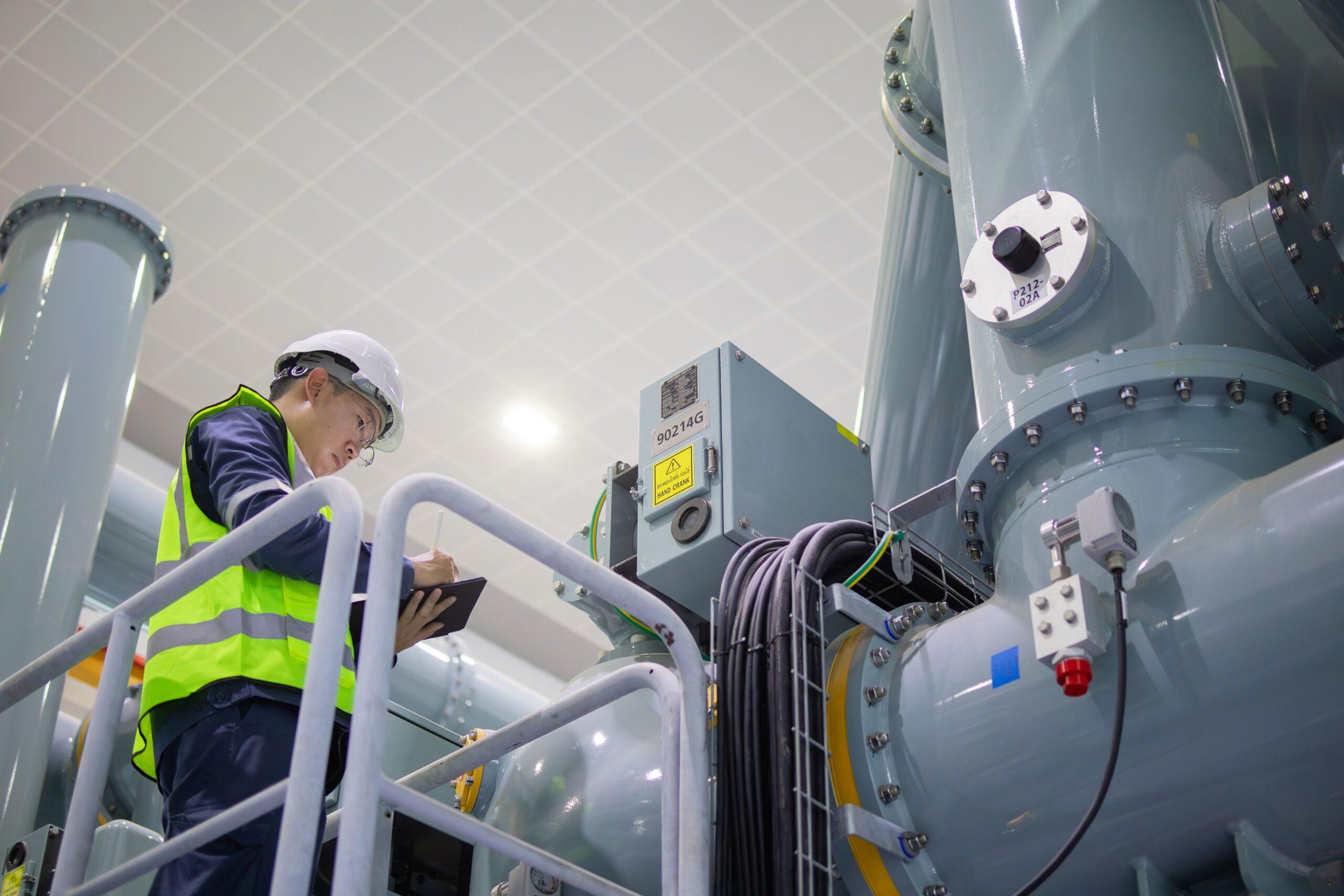 Techniker mit Schutzhelm und Warnweste kontrolliert elektrische Verbindungssysteme und notiert Prüfergebnisse in moderner Industrieanlage