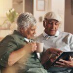 Aelteres Paar sitzt auf dem Sofa, nutzt ein Tablet und lacht gemeinsam – Symbol fuer digitale Unterstuetzung und Lebensfreude im Alltag beim Pflegedienst Bielefeld.